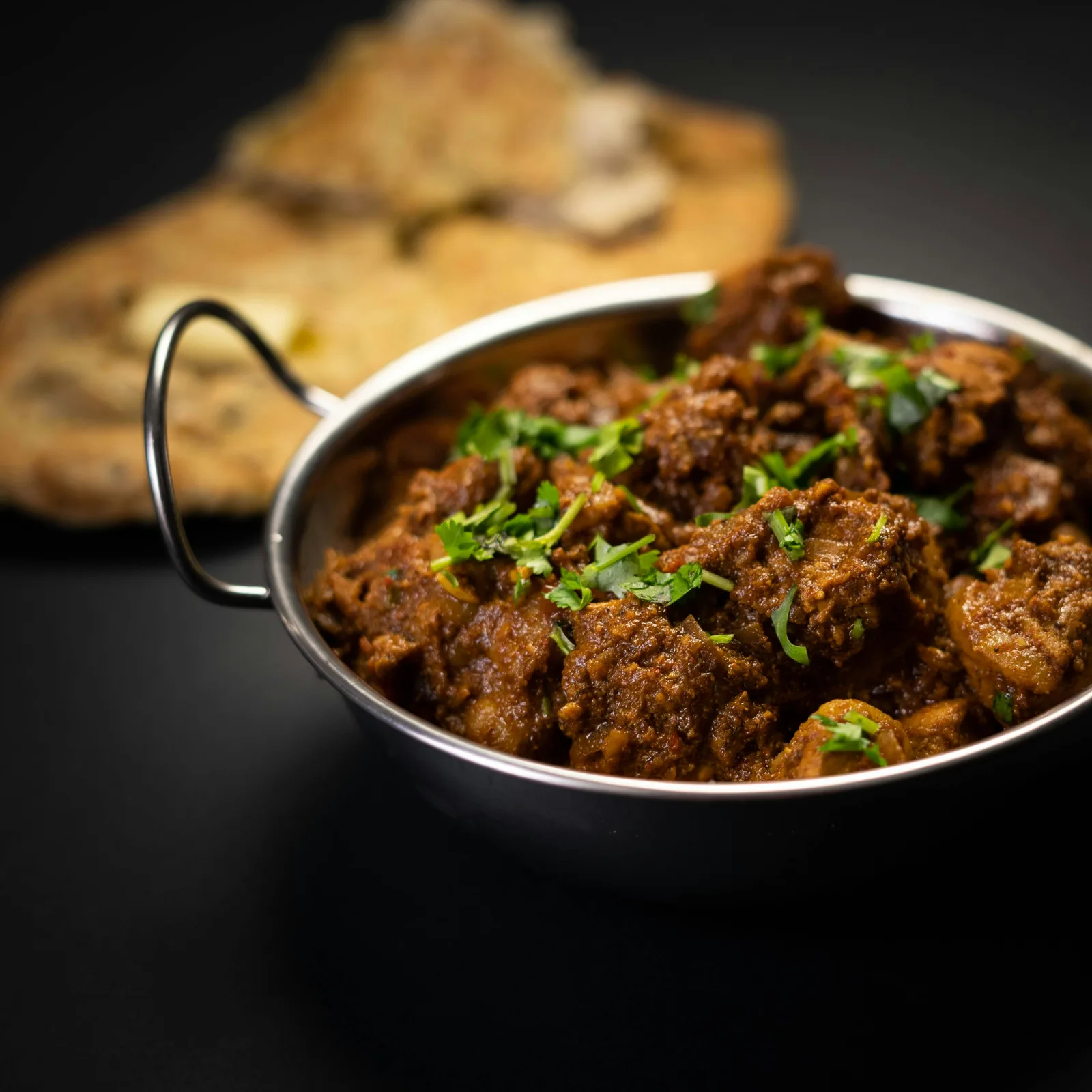 A copper pot of achar gosht, mutton curry in a rich red-brown gravy topped with green chillies, ginger and coriander, served with roti.
