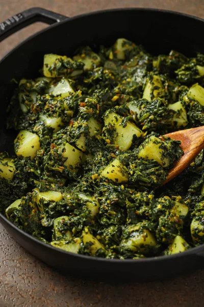 A bowl of Pakistani aloo palak with potato cubes in a thick green spinach gravy, served with roti and lemon wedges
