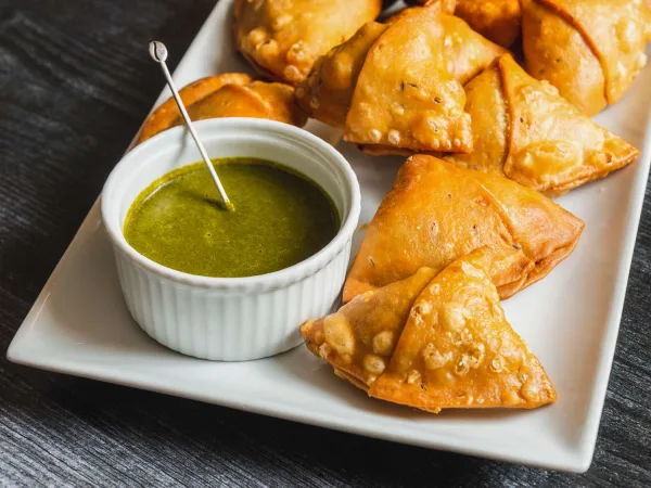 Golden fried aloo samosas on a plate, one broken open to show potato and pea filling, with chutneys beside them