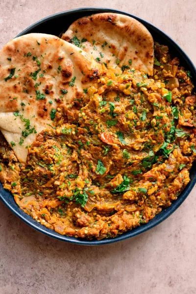 A pan of smoky Baingan Bharta garnished with coriander and ginger, served with roti and yogurt