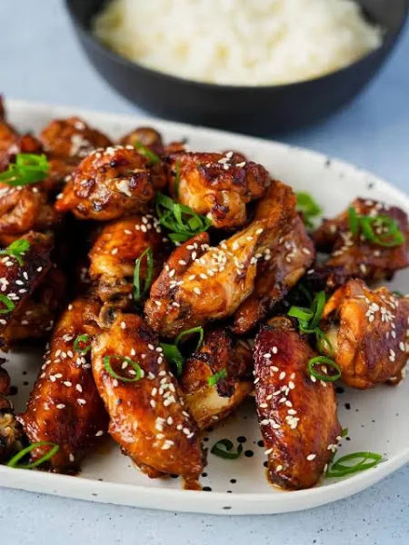 A platter of glossy grilled BBQ chicken wings with charred edges, garnished with fresh herbs and served with dipping sauce and vegetable sticks