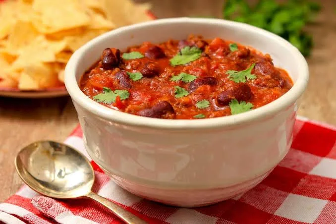 A bowl of thick beef chili with kidney beans, sour cream, cheddar and coriander next to a pot of chili and lime wedges