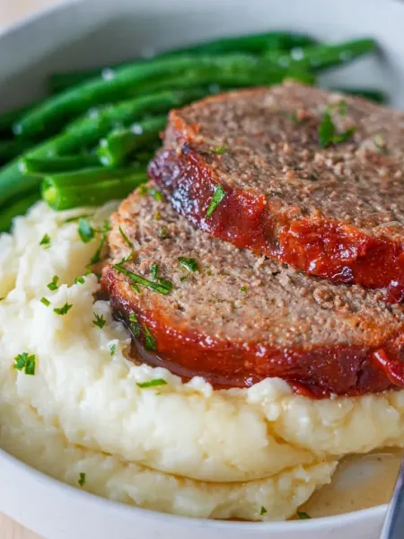 Slices of glazed beef meatloaf on a wooden board with mashed potatoes and green beans
