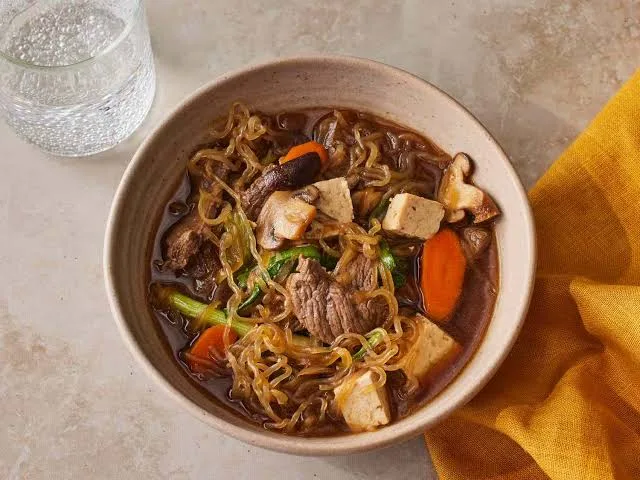Wide pan of beef sukiyaki with sliced beef, tofu, cabbage, mushrooms and noodles in a glossy soy broth, with rice and a bowl of beaten egg