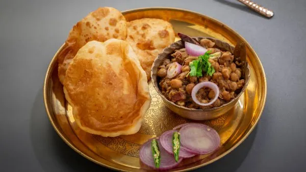 Golden puffed bhature on a steel plate with chole, onion rings, green chillies and lemon