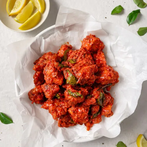 A plate of crispy red Chicken 65 with curry leaves, green chillies, onion rings and lemon wedges