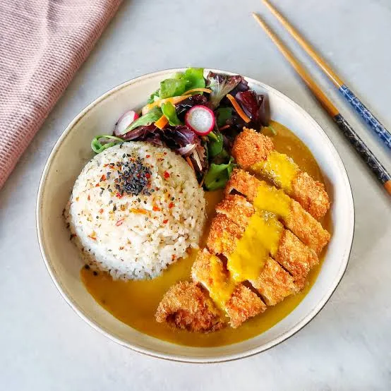 Sliced golden chicken katsu on rice with Japanese curry sauce and cucumber on a white plate