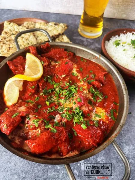 A pan of chicken tikka masala with charred chicken in a creamy orange tomato sauce, served with naan and lemon wedges