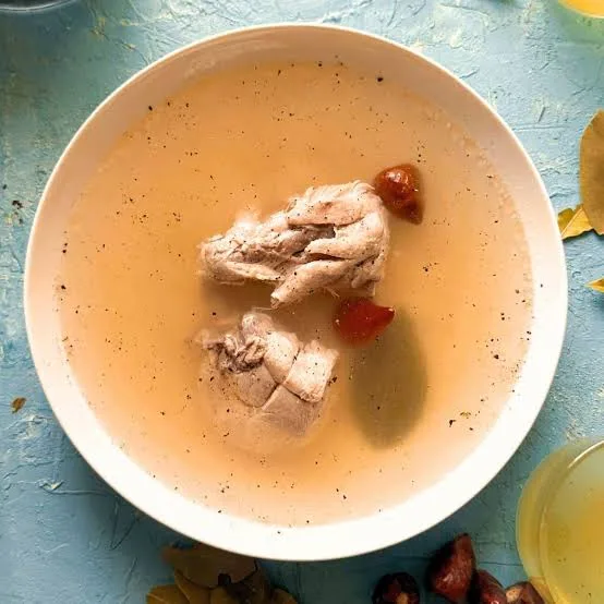 Bowl of clear golden chicken yakhni soup topped with shredded chicken, ginger and coriander, with lemon and whole spices beside it