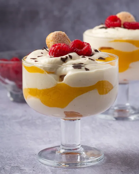 Glass bowl of custard trifle with layers of sponge, fruit, red jelly and custard under whipped cream and nuts