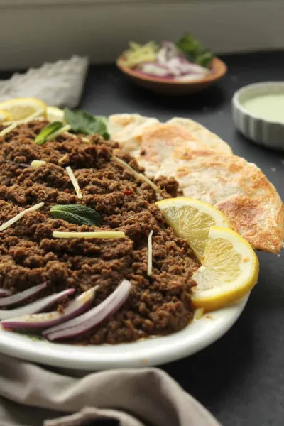 A pot of dum ka keema topped with fried onions, ginger and fresh herbs, served with naan and raita