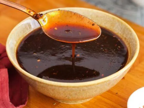 Bowl of glossy dark brown imli chutney with a spoon, surrounded by tamarind pods, jaggery and spices