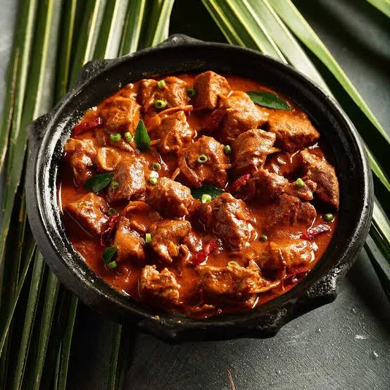 A pot of deep red rogan josh with tender lamb pieces, garnished with fresh coriander, beside rice and naan