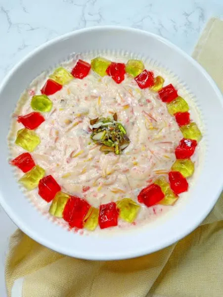 Glass bowl of Lab-e-Shireen with custard, red jelly cubes, mixed fruit, pistachios and pomegranate seeds