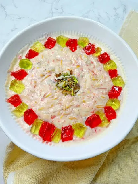 Glass bowl of Lab-e-Shireen with custard, red jelly cubes, mixed fruit, pistachios and pomegranate seeds