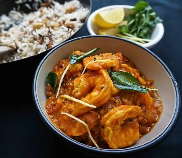 A pan of Malabar prawn curry in a rich orange coconut sauce with curry leaves, green chillies and coriander, served beside white rice.