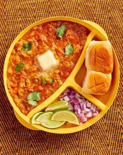 A bowl of buttery red pav bhaji topped with onion and coriander, served with toasted pav and lemon wedges