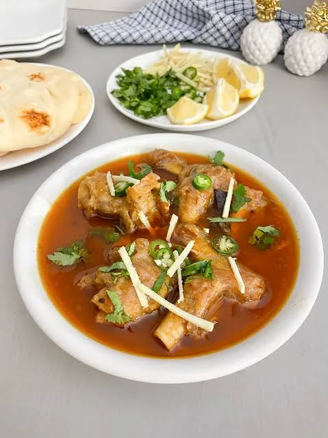 Bowl of Pakistani paya with tender trotters in a glossy spiced gravy, garnished with ginger, green chillies and coriander, served with naan and lemon.