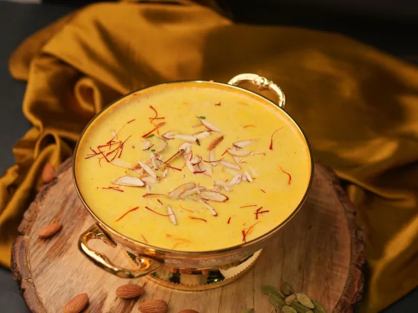 Soft white rasmalai discs in saffron milk topped with sliced pistachios in a white dish