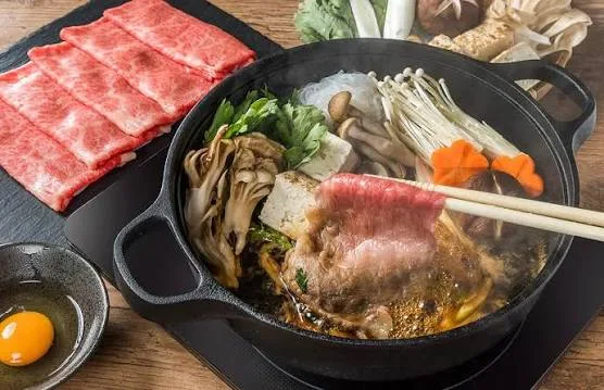 Shabu shabu hot pot with sliced beef, vegetables, tofu and two dipping sauces on a wooden table