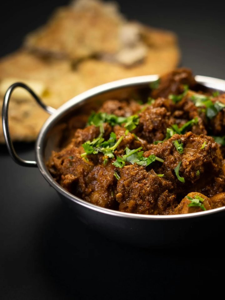 A copper pot of achar gosht, mutton curry in a rich red-brown gravy topped with green chillies, ginger and coriander, served with roti.