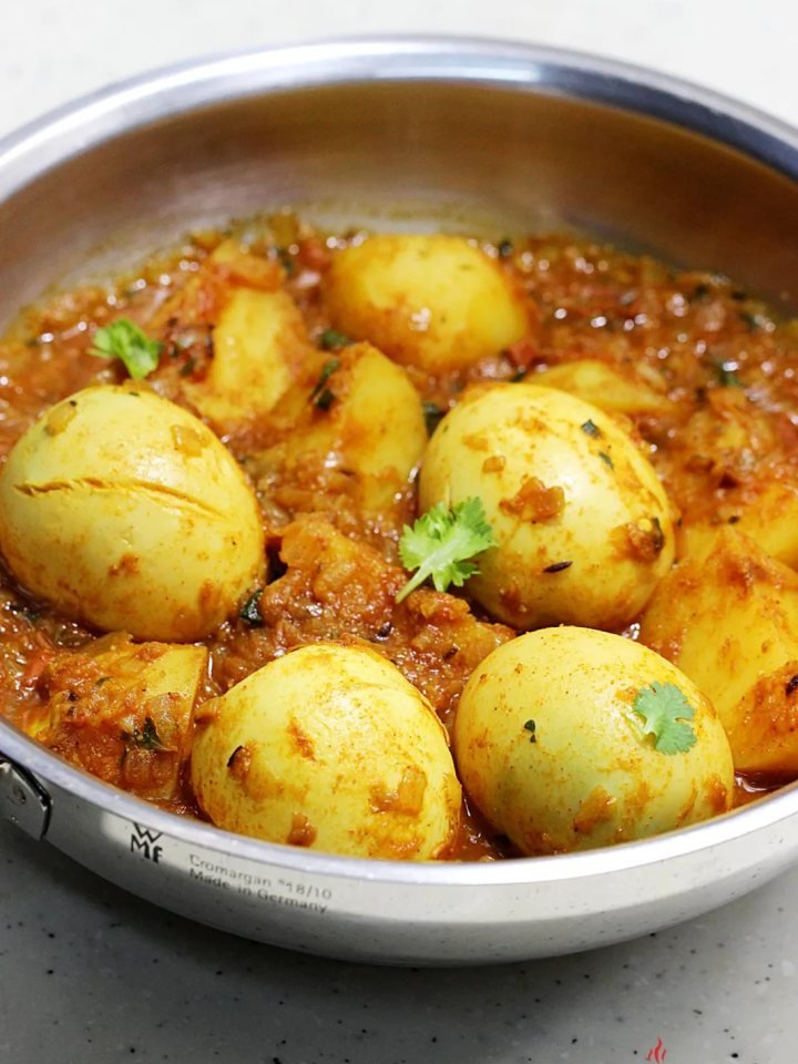 A pan of aloo anday with potato chunks and golden boiled eggs in red tomato gravy, garnished with green chillies and coriander