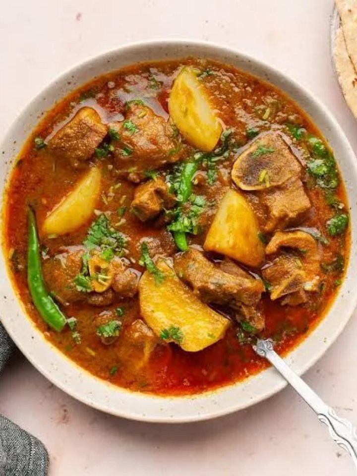 A pot of Pakistani aloo gosht with mutton pieces and potatoes in a rich red-brown gravy, garnished with coriander and ginger, served with roti.