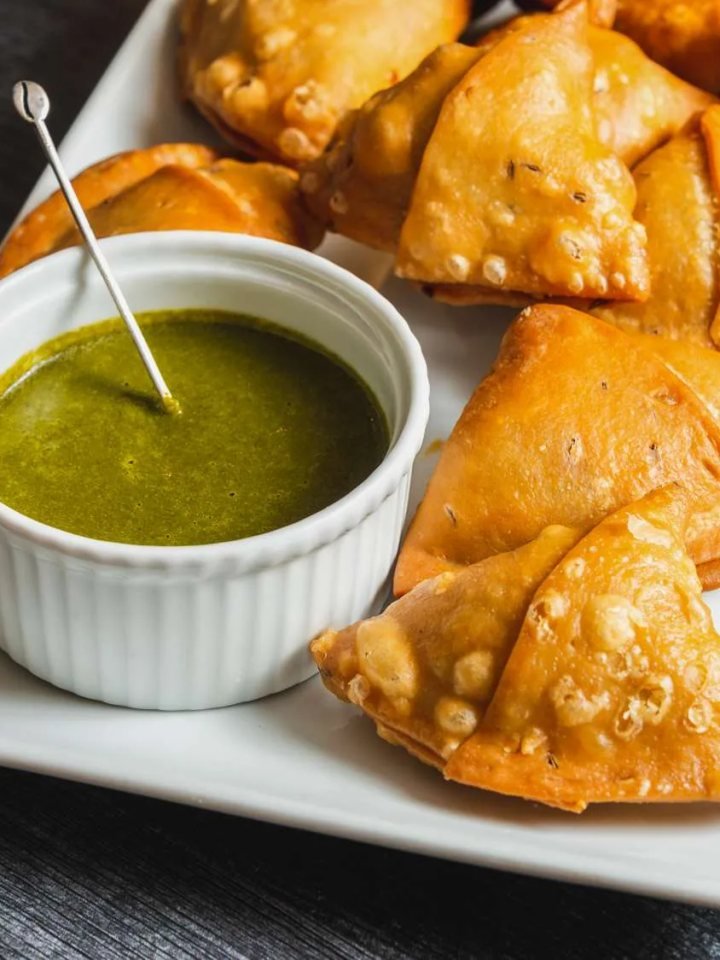 Golden fried aloo samosas on a plate, one broken open to show potato and pea filling, with chutneys beside them