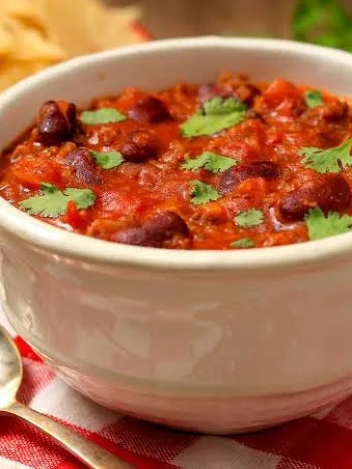 A bowl of thick beef chili with kidney beans, sour cream, cheddar and coriander next to a pot of chili and lime wedges