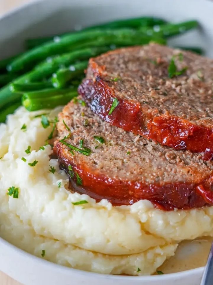 Slices of glazed beef meatloaf on a wooden board with mashed potatoes and green beans