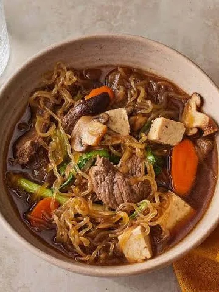 Wide pan of beef sukiyaki with sliced beef, tofu, cabbage, mushrooms and noodles in a glossy soy broth, with rice and a bowl of beaten egg