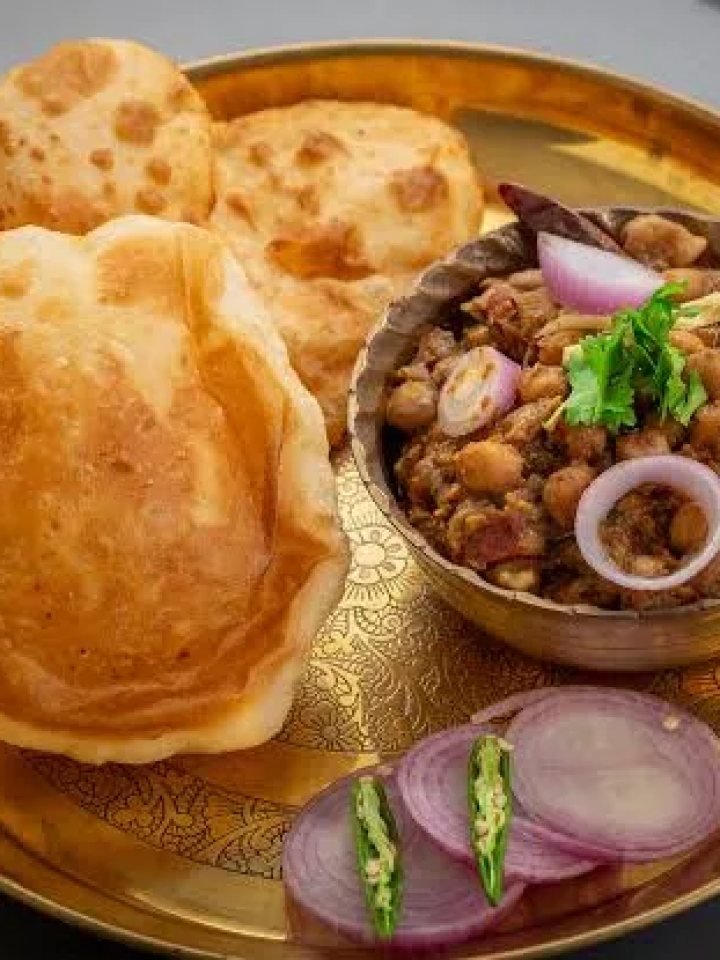 Golden puffed bhature on a steel plate with chole, onion rings, green chillies and lemon