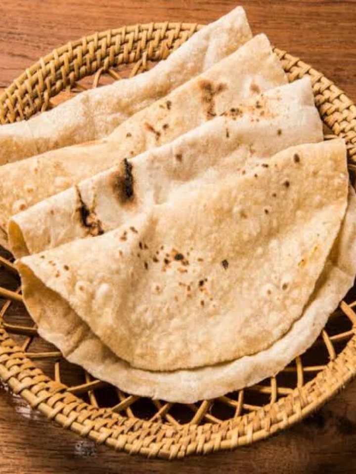 Stack of freshly cooked soft chapatis with golden spots on a wooden board beside a bowl of ghee