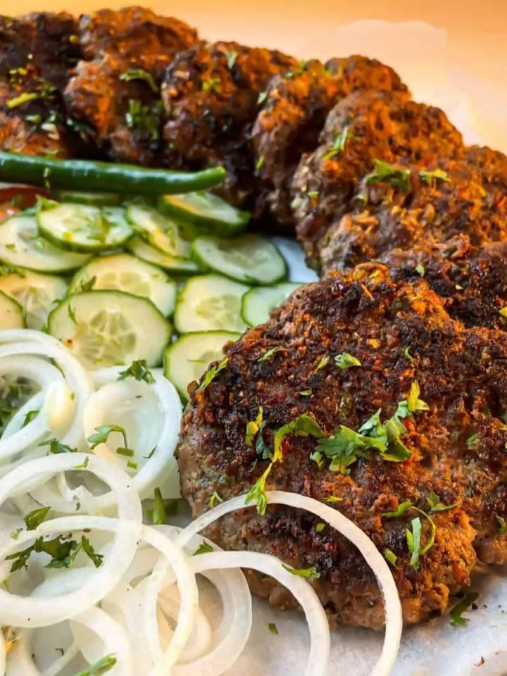 Golden brown chapli kababs on a slate board with naan, onion rings, lemon wedges and green chutney