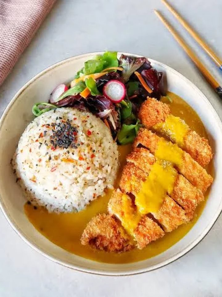 Sliced golden chicken katsu on rice with Japanese curry sauce and cucumber on a white plate