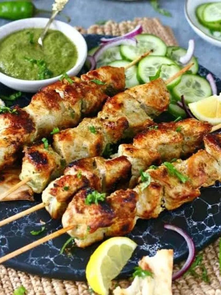 Skewers of creamy grilled chicken malai boti with lemon wedges, onion rings and green chutney on a slate board