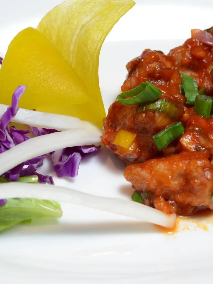 A white bowl of glossy chicken Manchurian with capsicum, onion and spring onion, served next to fried rice