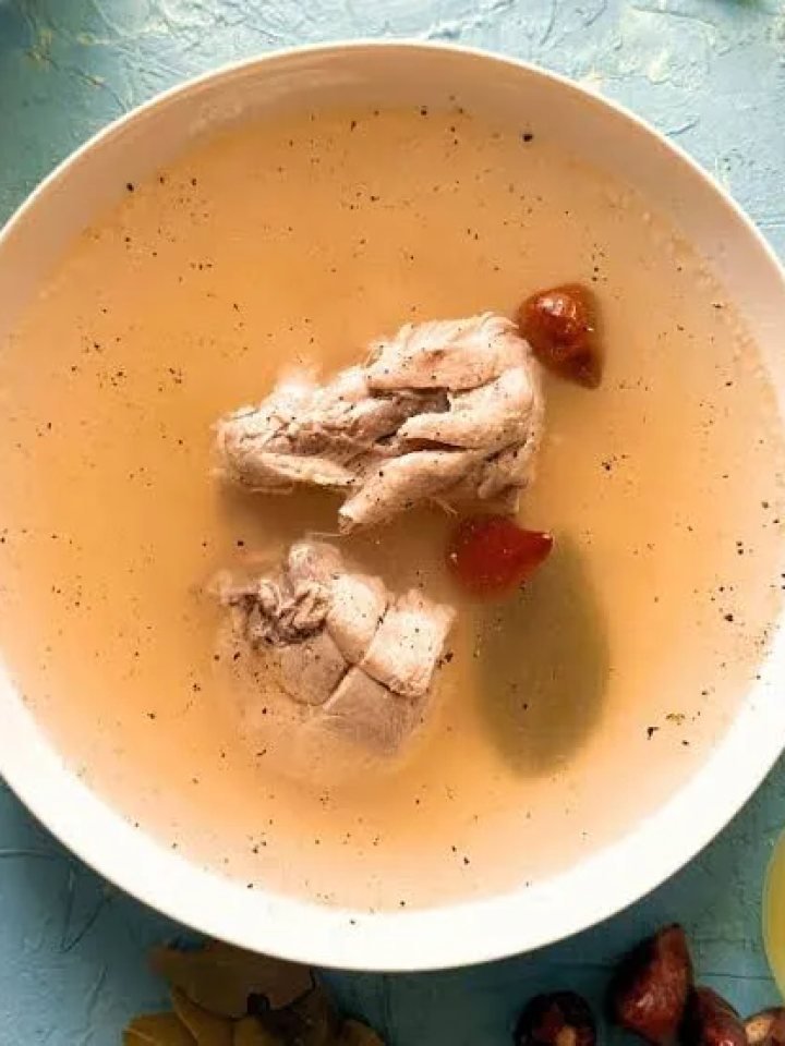Bowl of clear golden chicken yakhni soup topped with shredded chicken, ginger and coriander, with lemon and whole spices beside it