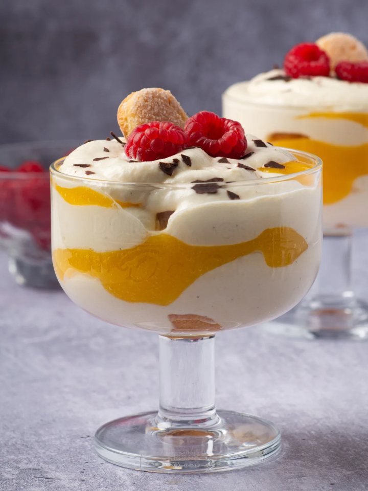 Glass bowl of custard trifle with layers of sponge, fruit, red jelly and custard under whipped cream and nuts