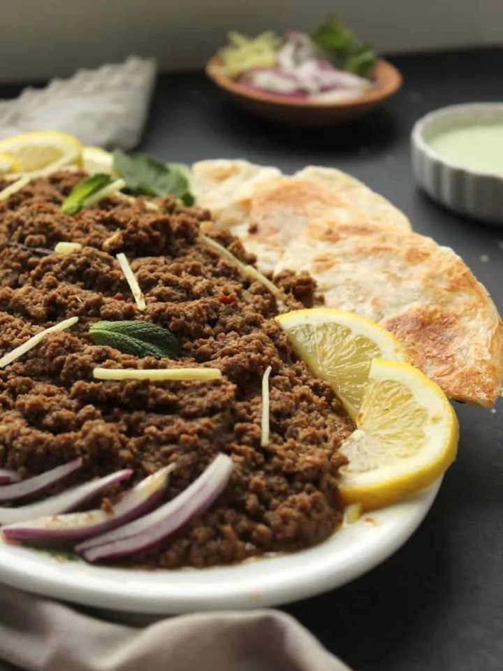 A pot of dum ka keema topped with fried onions, ginger and fresh herbs, served with naan and raita