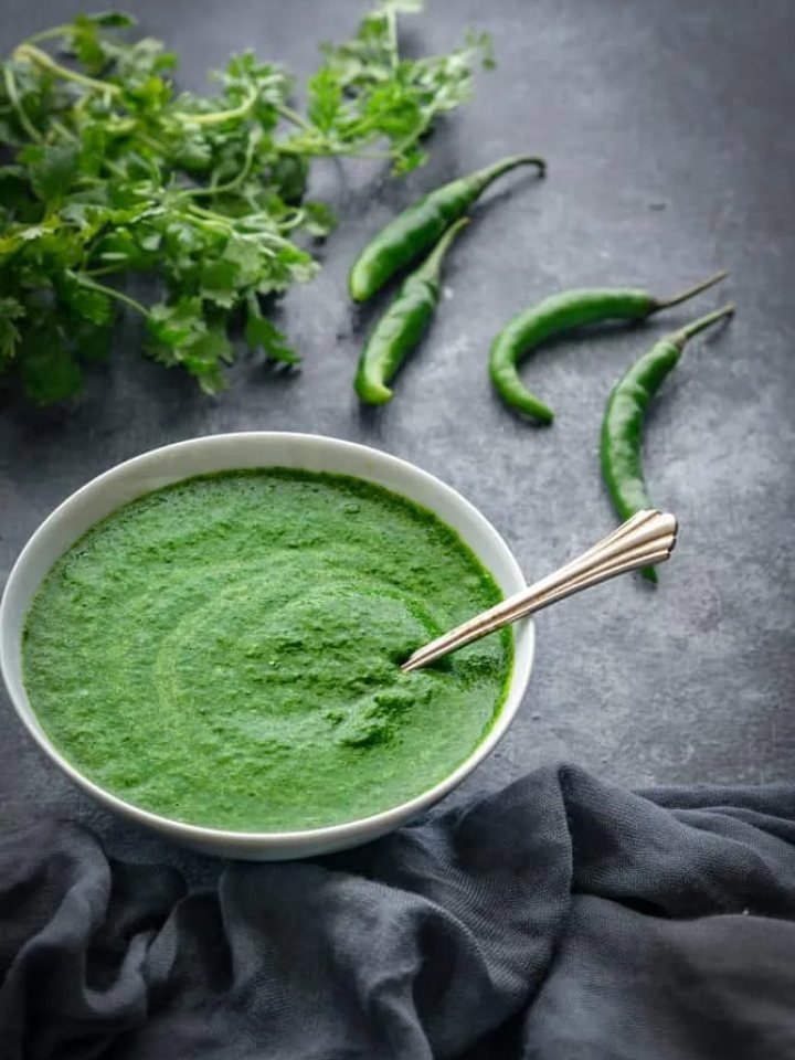 Bowl of bright green hari chutney with fresh coriander, green chillies and lemon on a wooden board