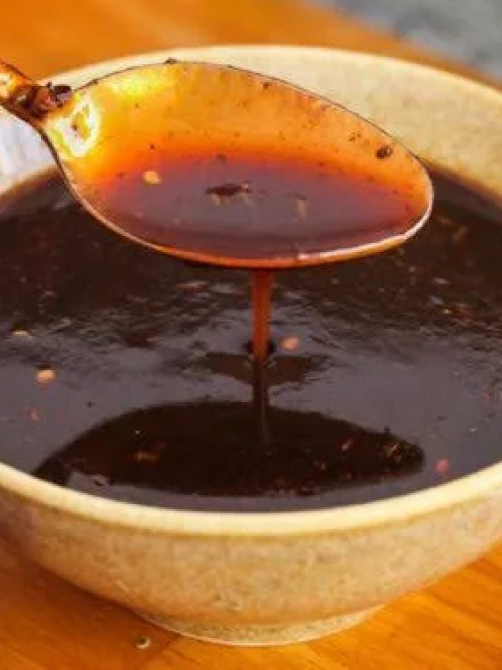 Bowl of glossy dark brown imli chutney with a spoon, surrounded by tamarind pods, jaggery and spices
