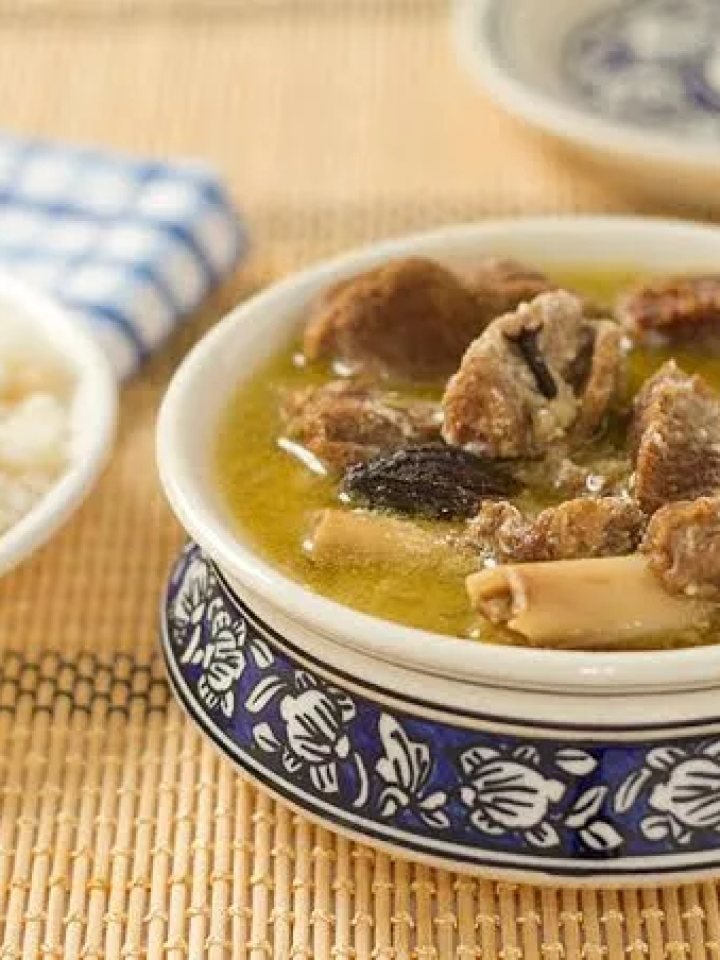 A pot of creamy pale Kashmiri yakhni with mutton pieces, whole spices and a bowl of white rice on a wooden table