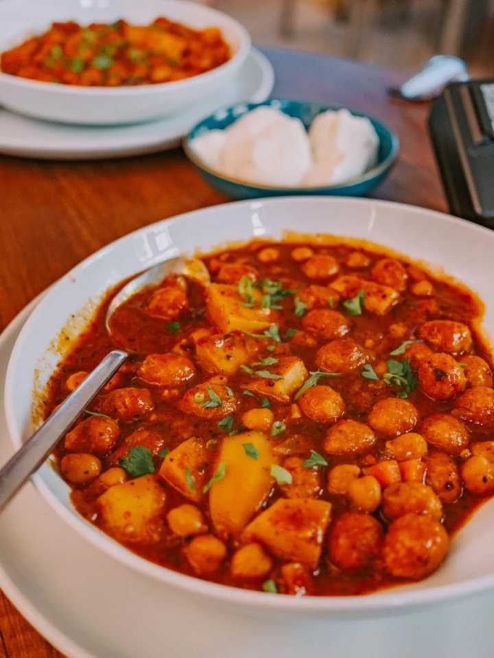 A brass bowl of dark Lahori chickpea curry topped with ginger and coriander, served with puri and lemon wedges