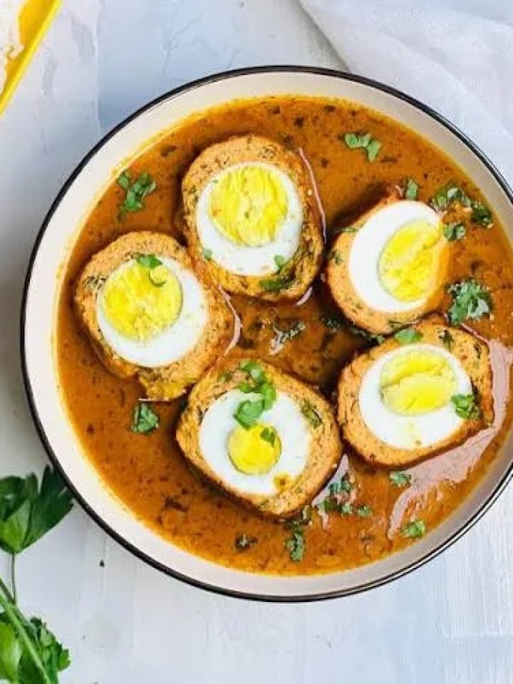 Halved nargisi koftas showing boiled egg centres in a glossy orange-brown gravy, garnished with green chillies and ginger