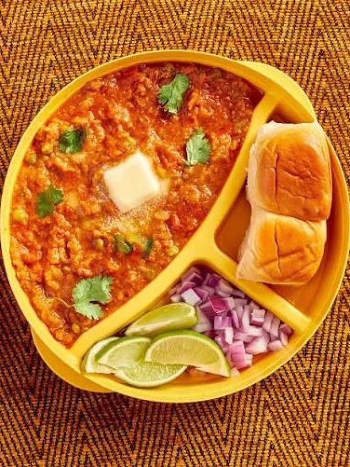 A bowl of buttery red pav bhaji topped with onion and coriander, served with toasted pav and lemon wedges
