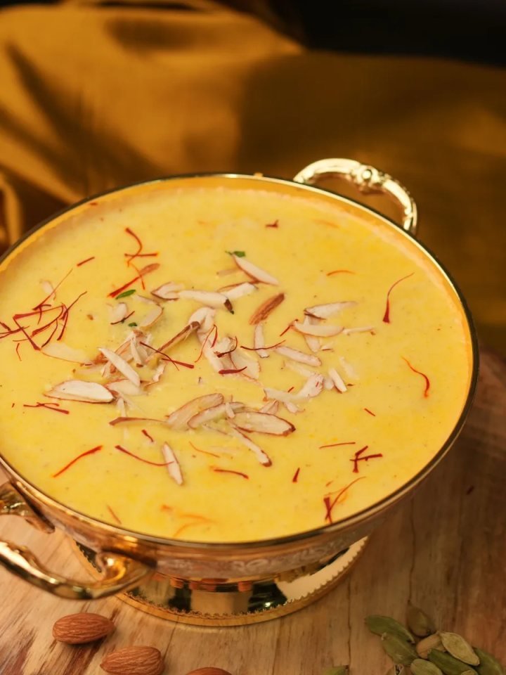 Soft white rasmalai discs in saffron milk topped with sliced pistachios in a white dish