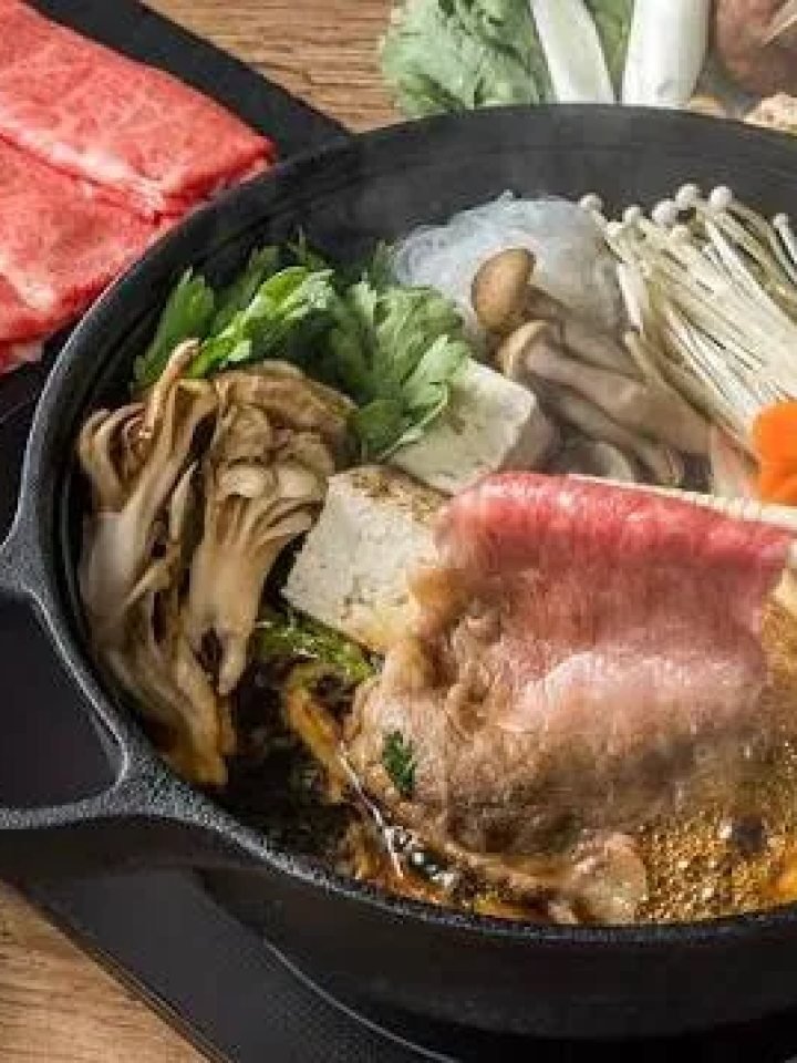 Shabu shabu hot pot with sliced beef, vegetables, tofu and two dipping sauces on a wooden table