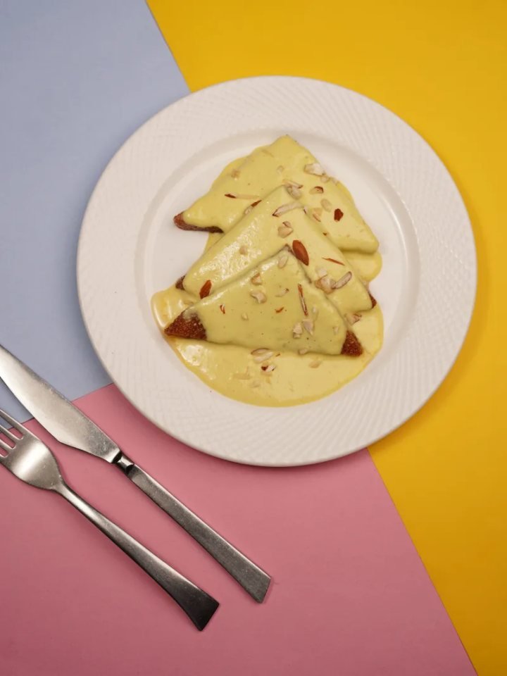 Golden fried bread triangles topped with thick saffron milk and sliced pistachios in a white dish