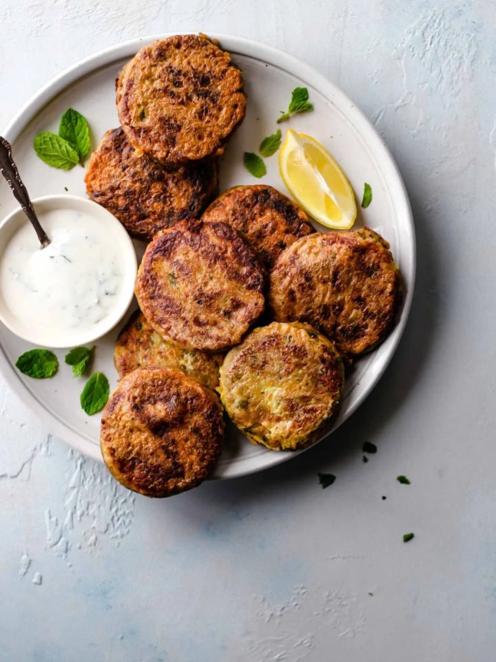 Golden shallow-fried shami kababs on a plate with green chutney, lemon wedges and sliced onions